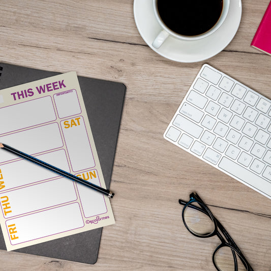 A weekly planner sheet with a 'THIS WEEK' layout, placed on a wooden desk alongside a keyboard, coffee, glasses, and a pencil.