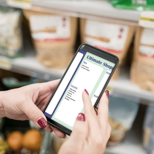A person holding a smartphone in a grocery store aisle, with the phone screen showing a shopping list app under the heading 'Ultimate Shopping List'.