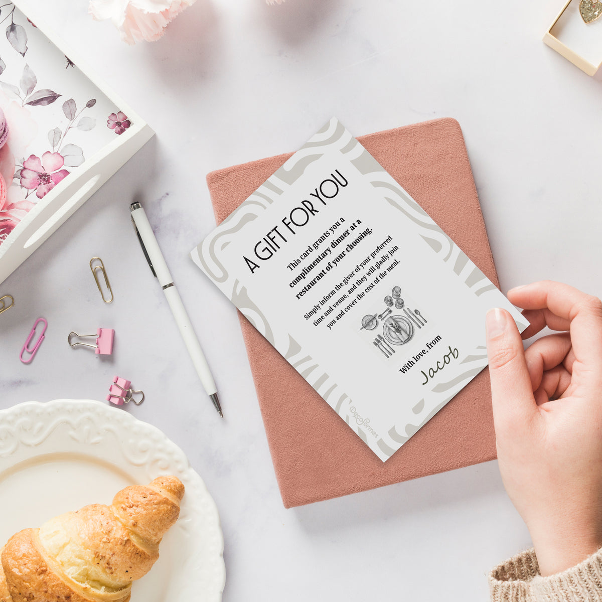 A hand holds a white 'A Gift for You' complimentary dinner card over a dusty rose notebook, arranged in a styled flat-lay with a croissant, stationery, and a floral box on a white marble surface.